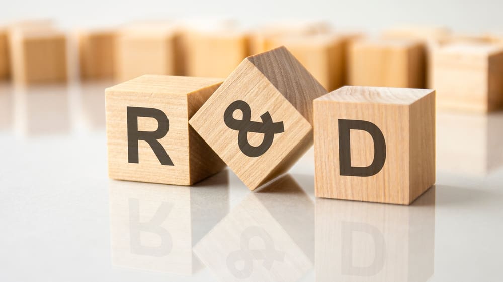 Wooden blocks with the letters "R," "&," and "D" placed on a white surface, representing "Research and Development." Blurry blocks are in the background.