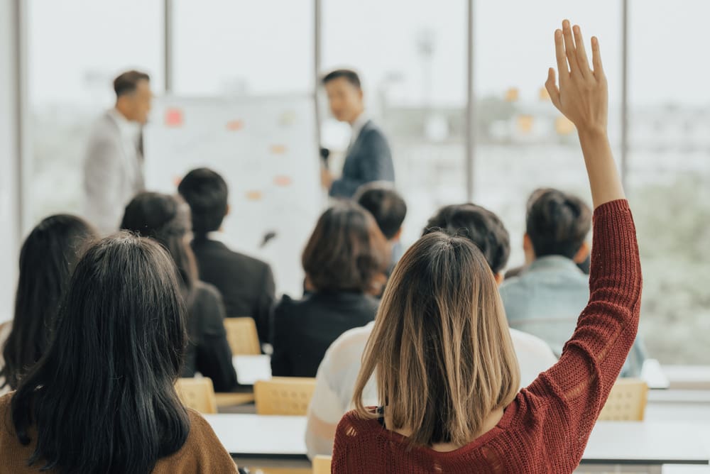 A group of people are attending a presentation in a classroom or seminar setting. One person in the foreground, wearing a red sweater, is raising their hand to ask a question about the difference between R&D tax credits and R&D allowance. The presenter at the front is pointing to a whiteboard with various notes or diagrams on it.